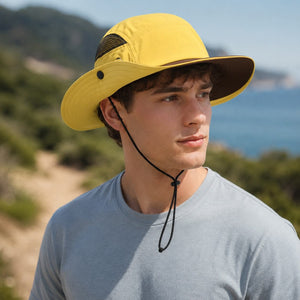 Male model wearing a yellow outdoor sun protection hat with wide brim and adjustable chin cord near a coastal hiking trail.