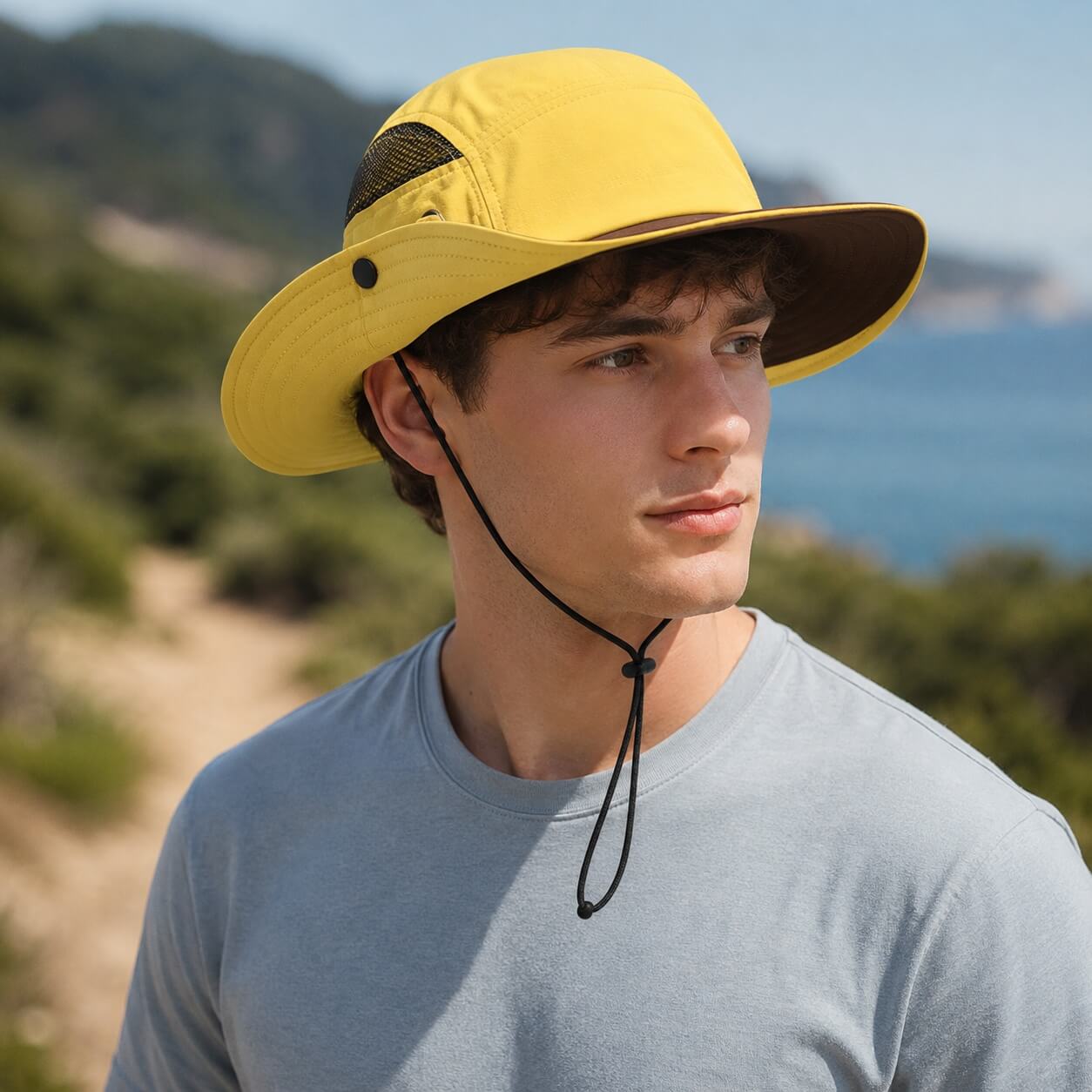 Male model wearing a yellow outdoor sun protection hat with wide brim and adjustable chin cord near a coastal hiking trail.
